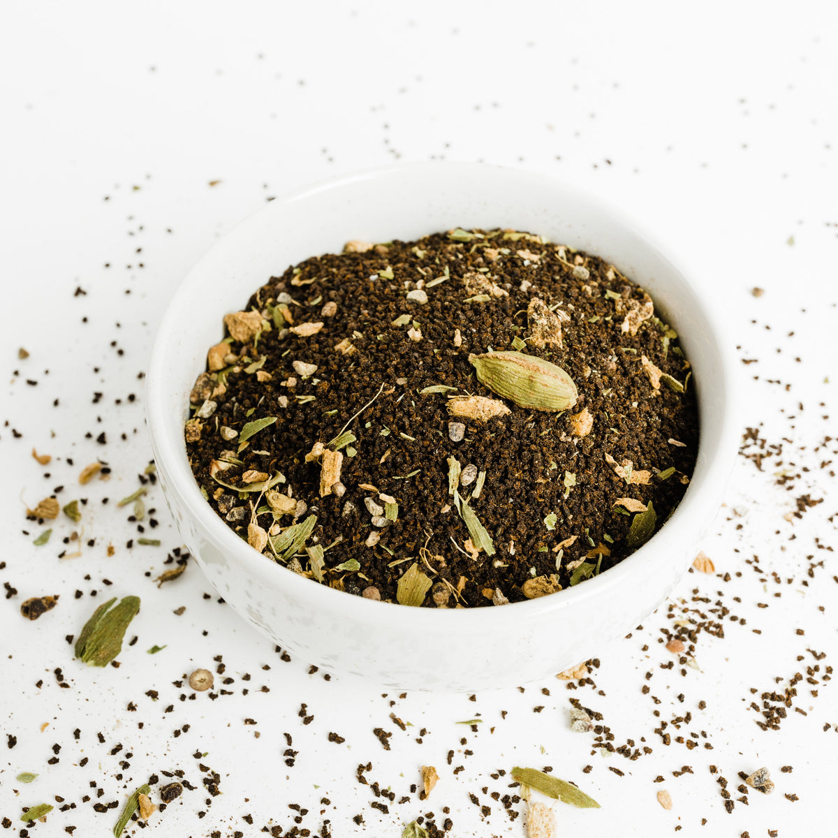 Loose Chai Me At Home blend in a white bowl showing dark CTC black tea granules, green cardamom pods and pieces of dried ginger