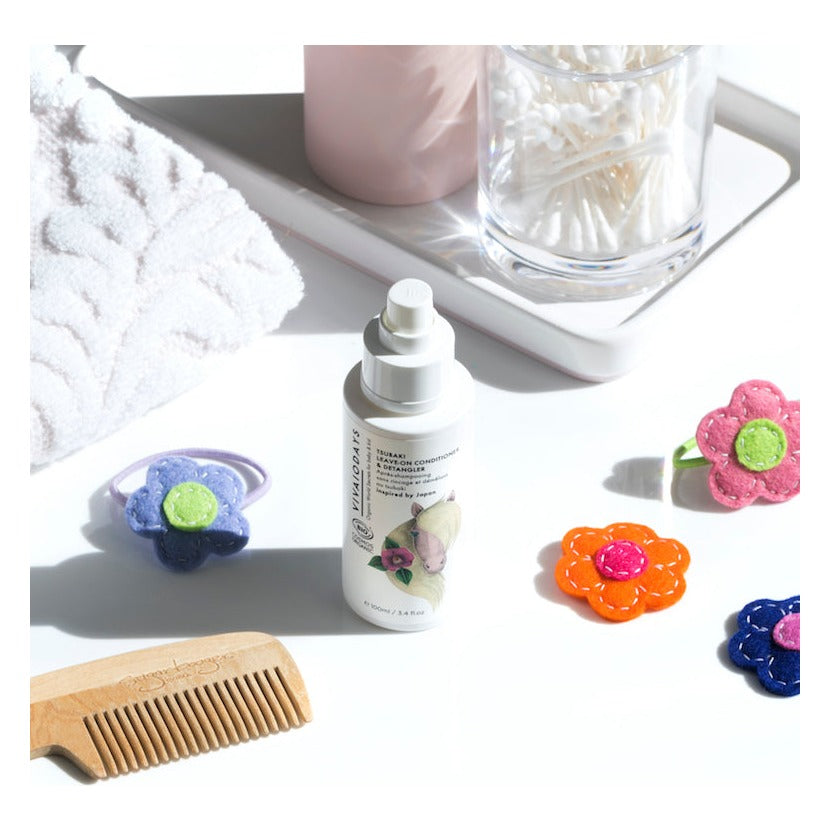 Vivaiodays Tsubaki Leave-In Conditioner & Detangler spray bottle on a white bathroom counter next to a wooden comb and felt flower hair clips