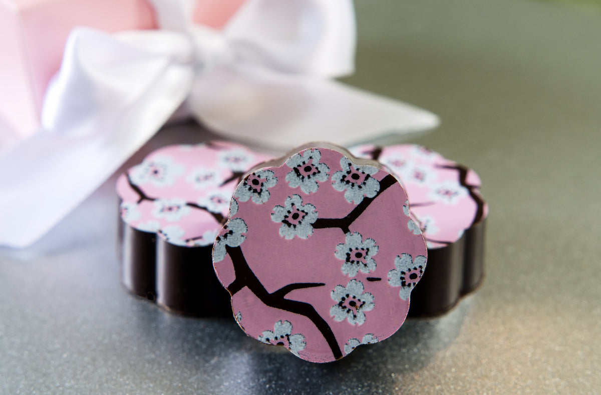 Chouquette Cherry Blossom chocolate caramels with pink tops printed with white blossoms and dark branches, in front of a white ribbon