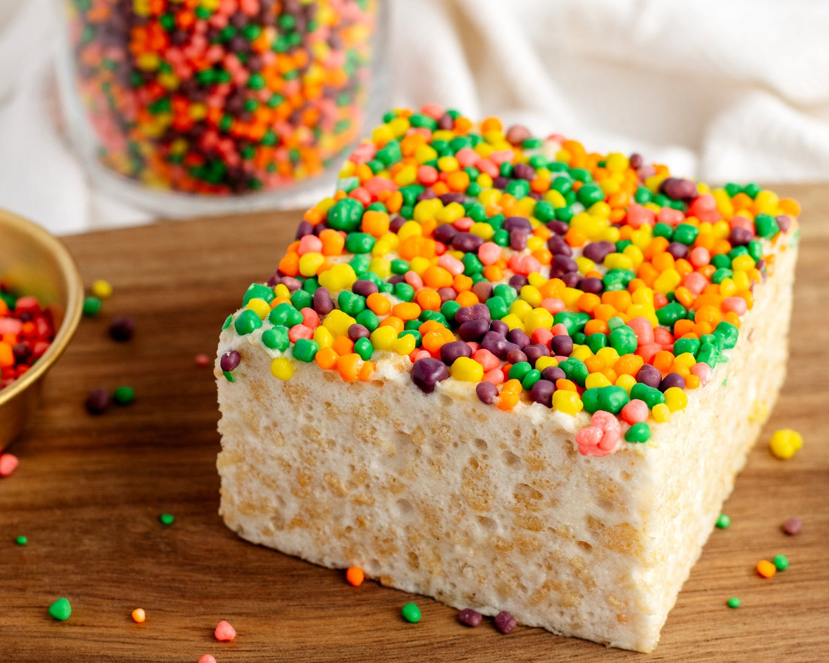 Close side view of the Nerdtastic Crispycake on a wooden board, showing the candy top and crispy rice layer, with a jar of candies behind