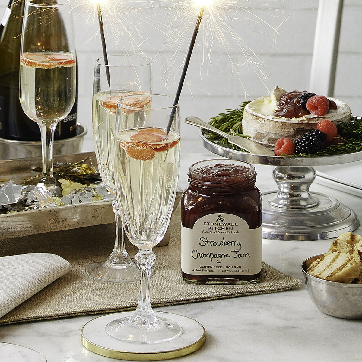Open jar of Stonewall Kitchen Strawberry Champagne Jam on a New Year's table with champagne flutes, sparklers and a cheese platter