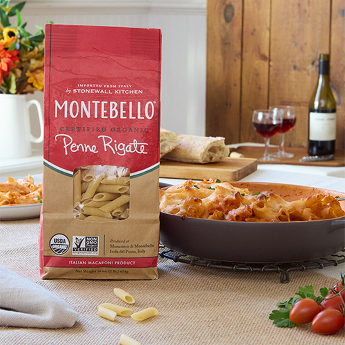 Bag of Montebello Organic Penne Rigate on a dining table next to a dish of baked penne, sliced bread and glasses of red wine