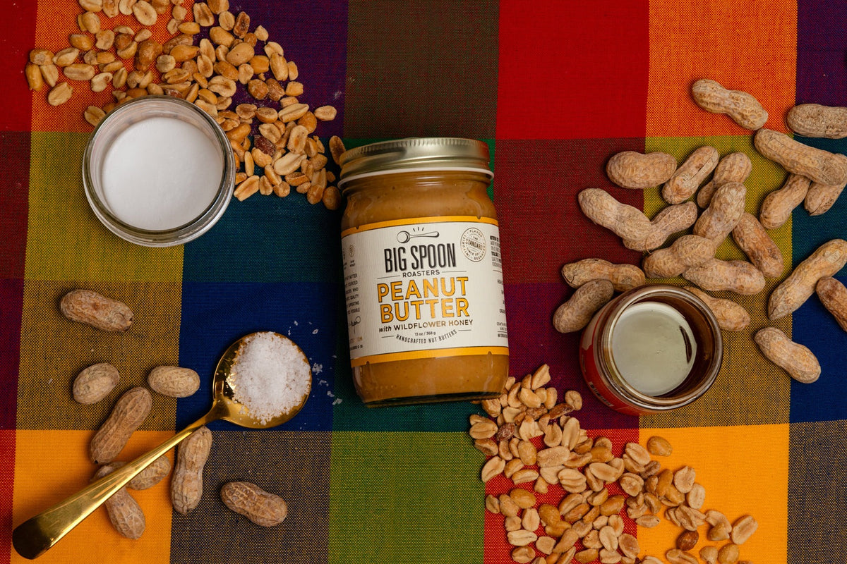 Jar of Big Spoon Roasters Peanut Butter with Wildflower Honey on a plaid cloth with shelled and in-shell peanuts, honey, coconut oil and a spoon of sea salt