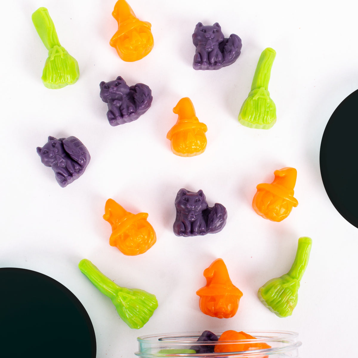 Witches' Brew gummies scattered on white: green broomsticks, purple cats and orange jack-o'-lanterns