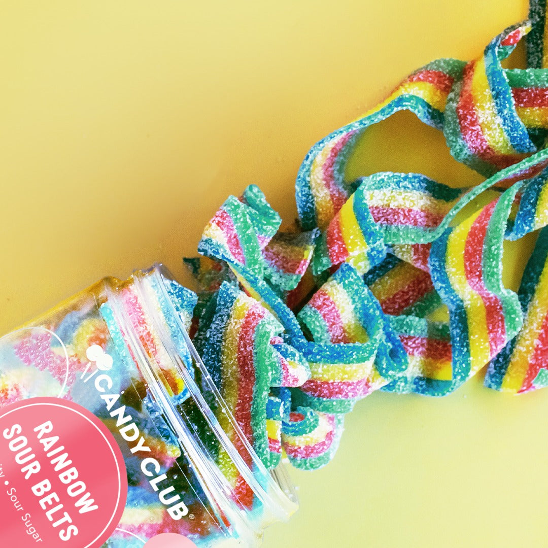 Rainbow-striped sour belts spilling from a tipped Candy Club Rainbow Sour Belts cup onto a yellow background