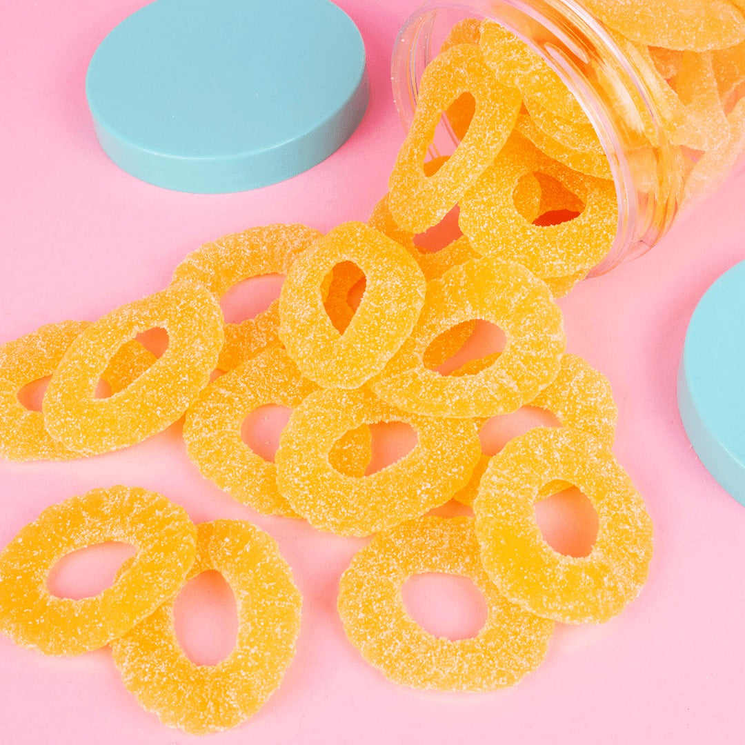 Sugar-coated Candy Club Pineapple Rings gummies spilling from a clear cup onto a pink surface beside teal lids