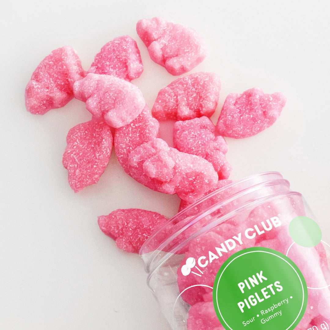 Candy Club Pink Piglets cup tipped over with pink pig-shaped sour gummies spilling onto a white surface