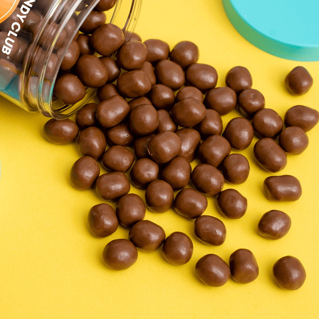 Candy Club Cookie Dough Bites spilling from a tipped cup onto a yellow surface, showing the round milk chocolate-covered pieces