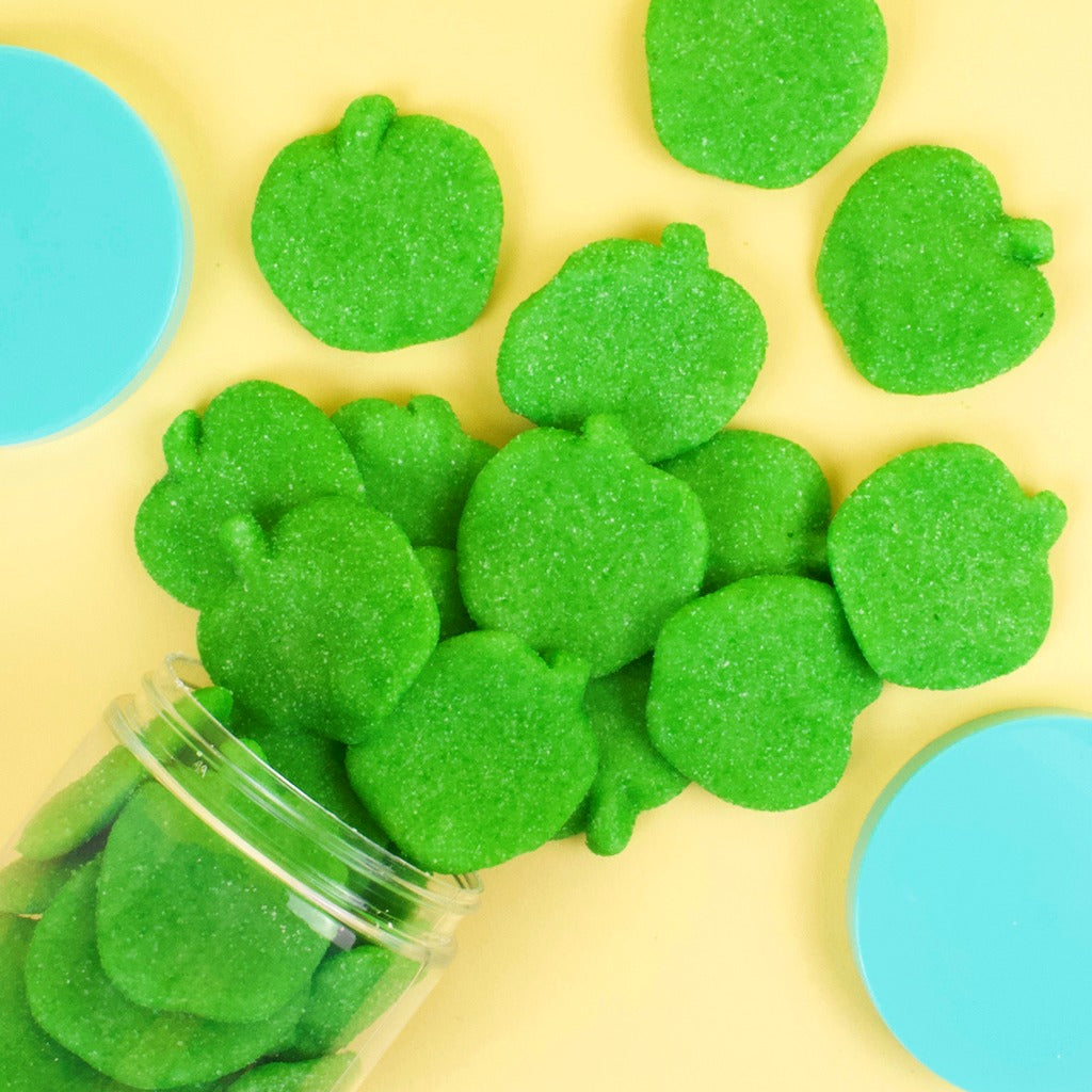 Candy Club Big Apples green apple-shaped sugar-sanded gummies spilling from a tipped cup onto a yellow surface with teal lids