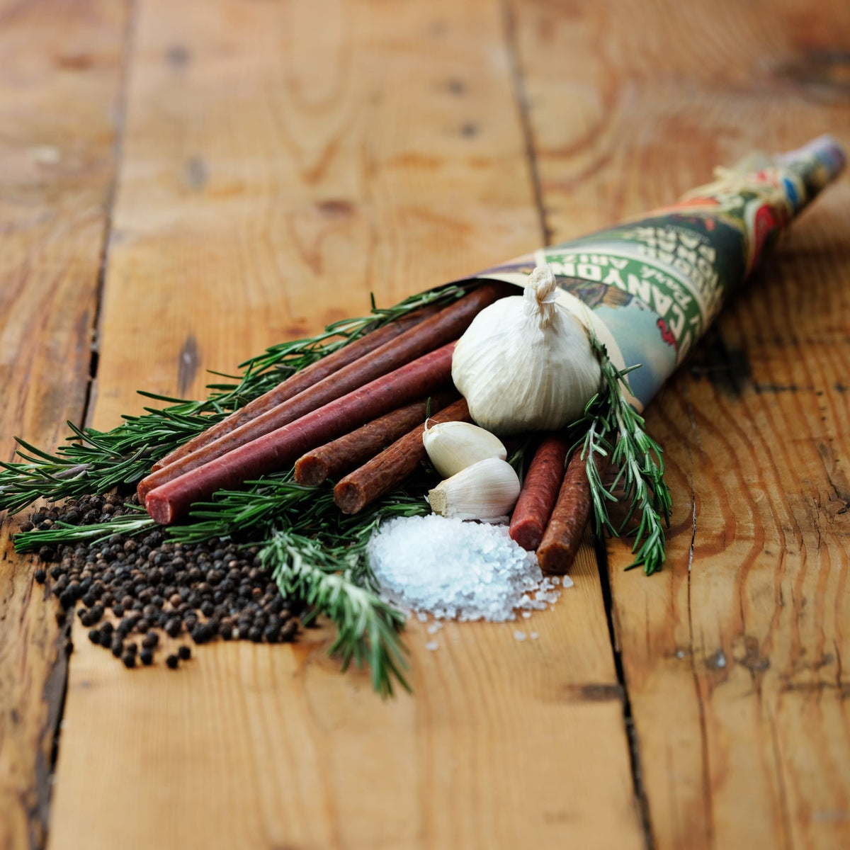 Field Trip Cracked Pepper Turkey Sticks on a wooden board with garlic, rosemary, black peppercorns and coarse salt