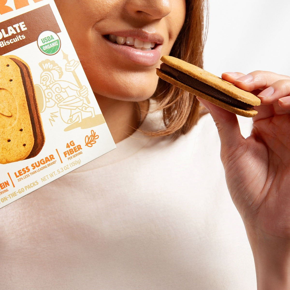 Person biting an Olyra dark chocolate crème sandwich biscuit, with the Dark Chocolate Crème Breakfast Biscuits box in the foreground