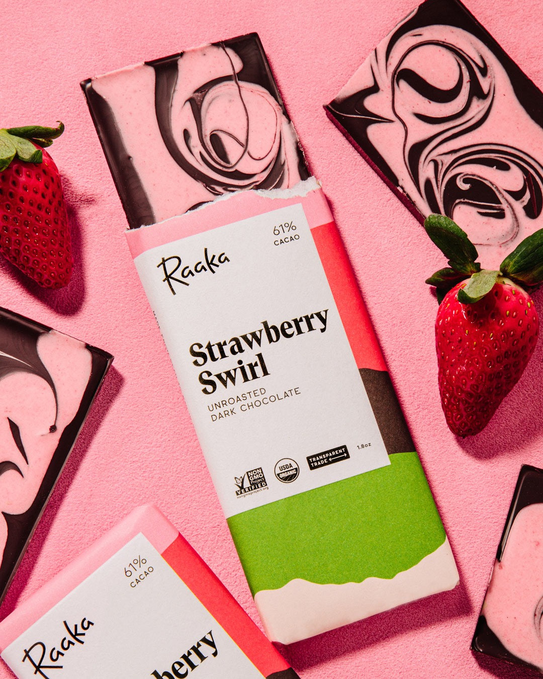 Raaka Strawberry Swirl 1.8 oz bars on a pink background with broken swirled pieces and fresh strawberries