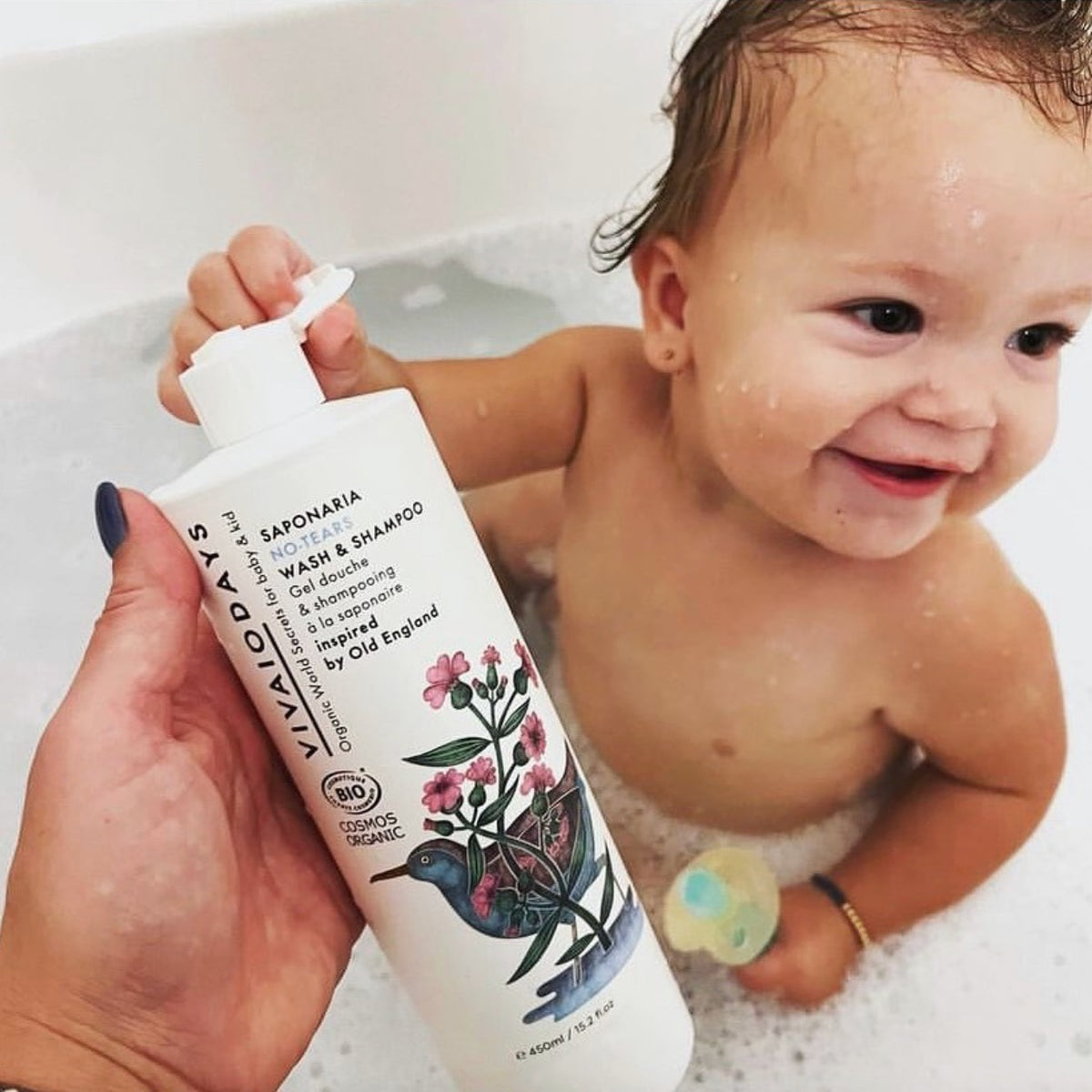Adult hand holding the Vivaiodays Saponaria No-Tears Wash & Shampoo 450 ml bottle beside a smiling baby in a bubble bath