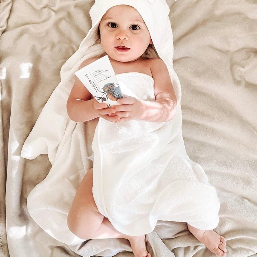 Baby wrapped in a white hooded towel holding a Vivaiodays Olive Oil Rescue Balm Gel tube
