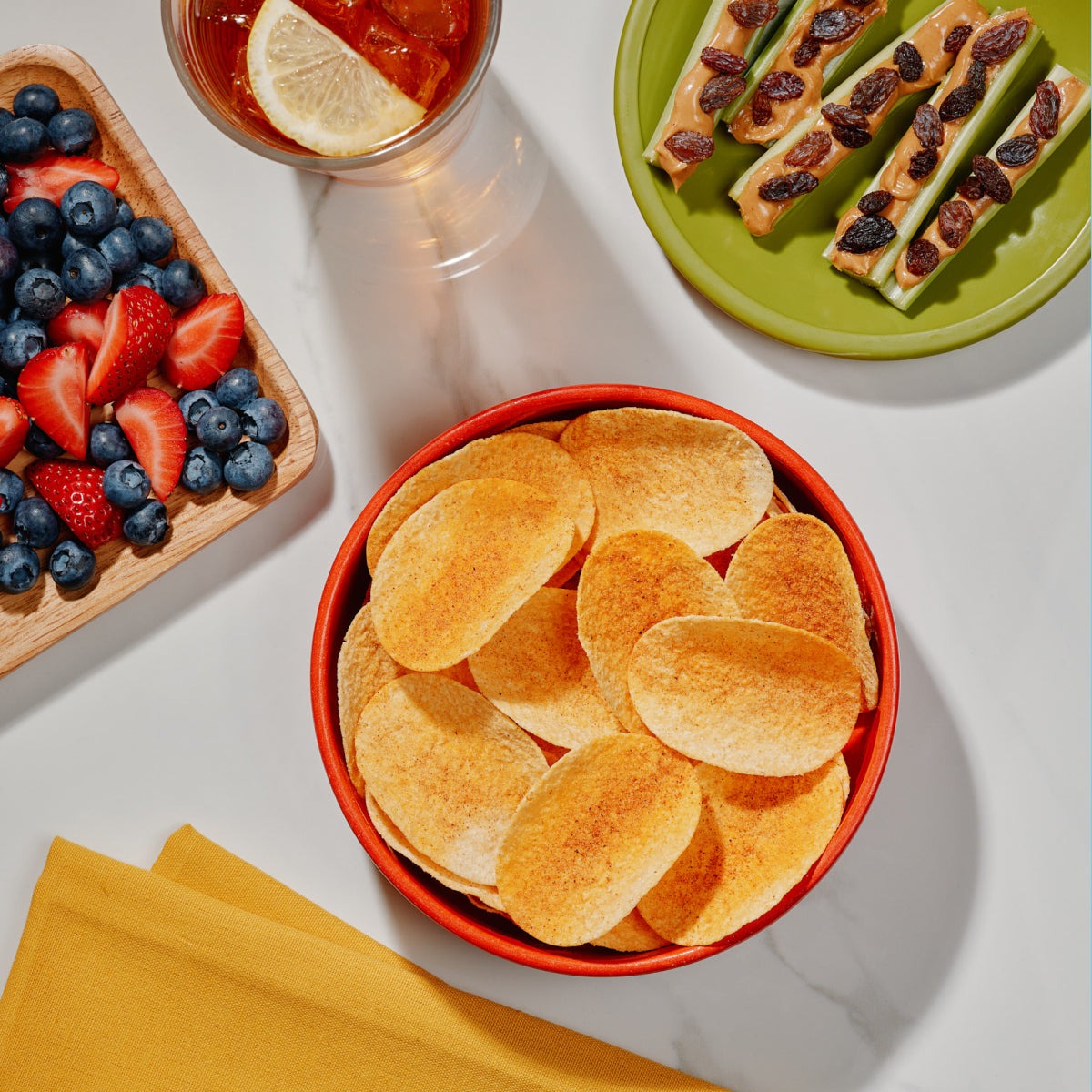 Bowl of Outback BBQ seasoned potato crisps on a white table beside berries, celery sticks and iced tea