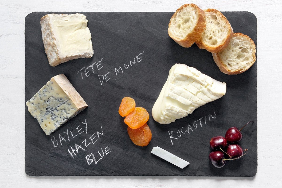 Black 10 x 14 inch Brooklyn Slate cheese board from above with three cheeses labeled in chalk, sliced baguette, dried apricots, cherries and a soapstone chalk stick