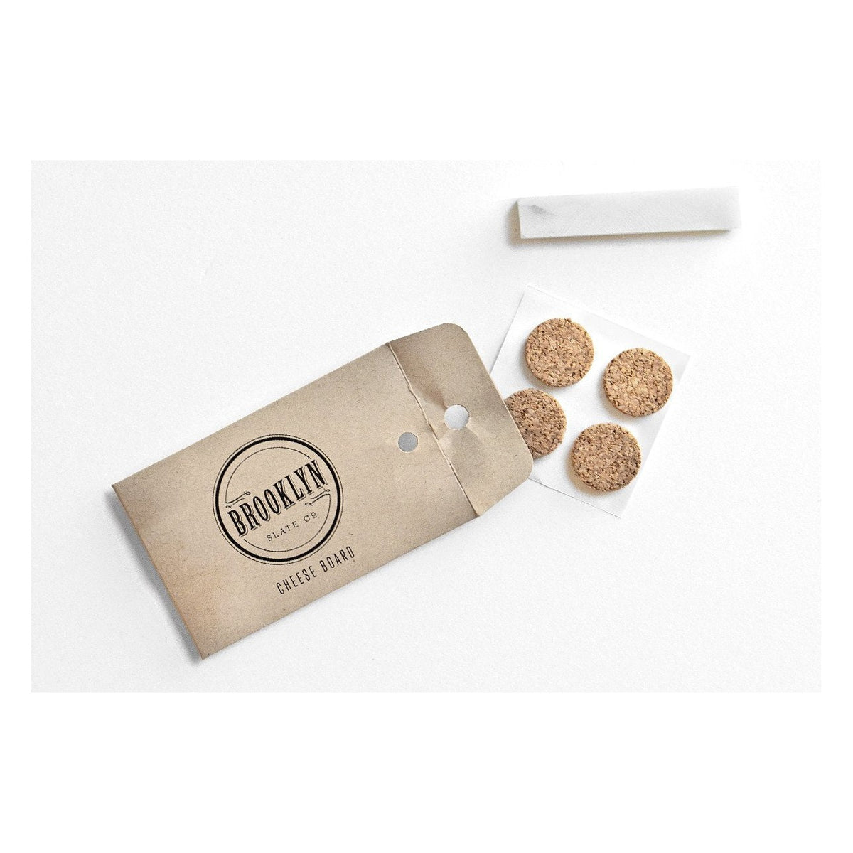 Brooklyn Slate Co. cheese board accessory envelope with a stick of white soapstone chalk and four round anti-slip feet that come with the board