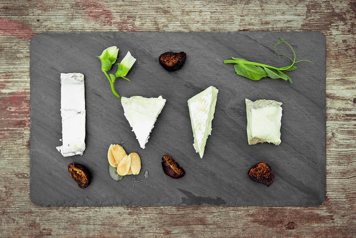 Black 7 x 12 inch Brooklyn Slate cheese board from above on a wooden table with four soft cheeses, dried figs, sliced almonds and pea shoots