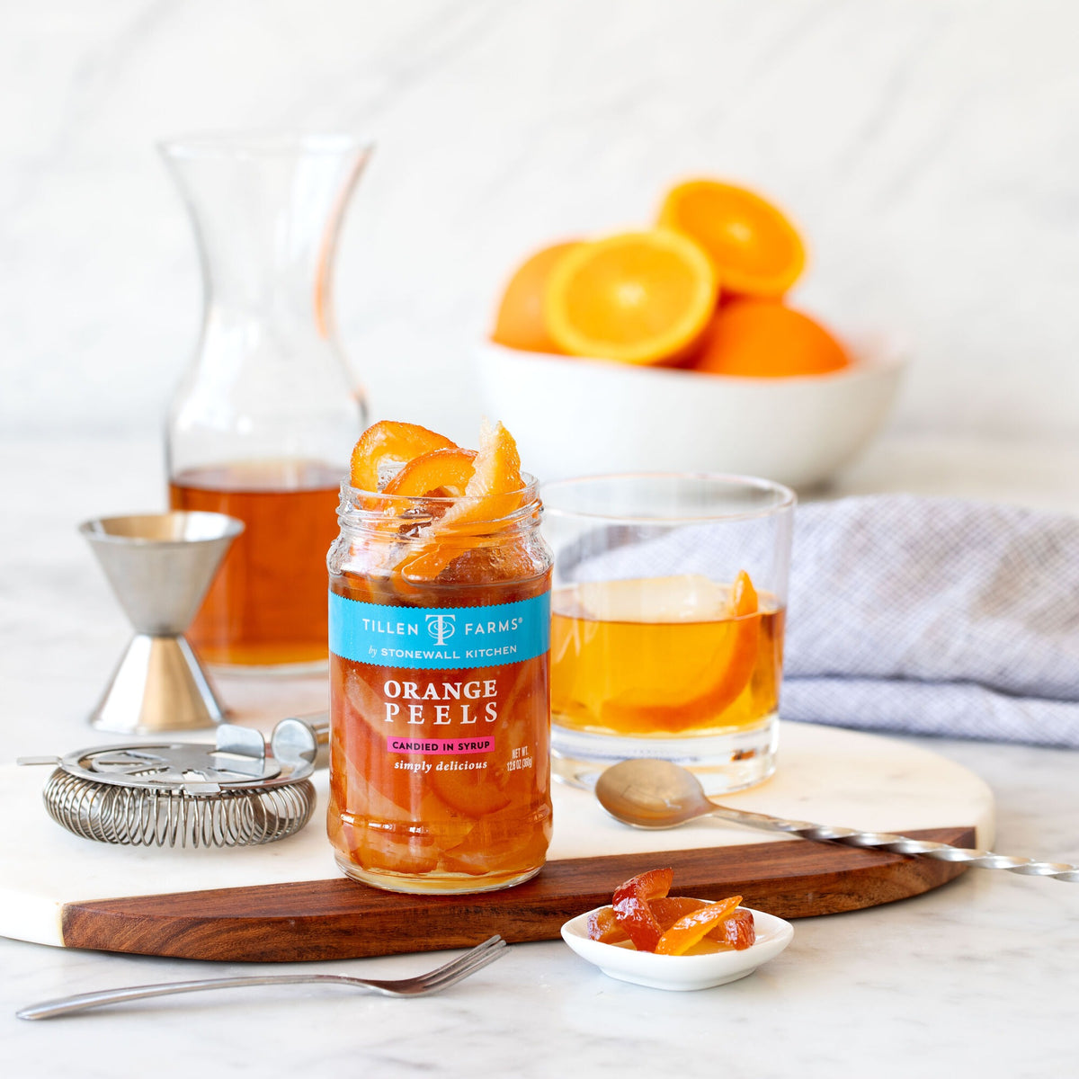Open jar of Tillen Farms Orange Peels Candied in Syrup on a wooden board beside an Old Fashioned cocktail, a bar strainer and a small dish of candied peel
