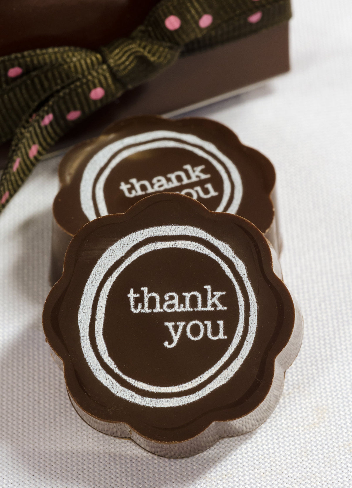 Two Chouquette Thank You! chocolates with white typewriter-style thank you lettering inside a double ring, beside a ribboned gift box