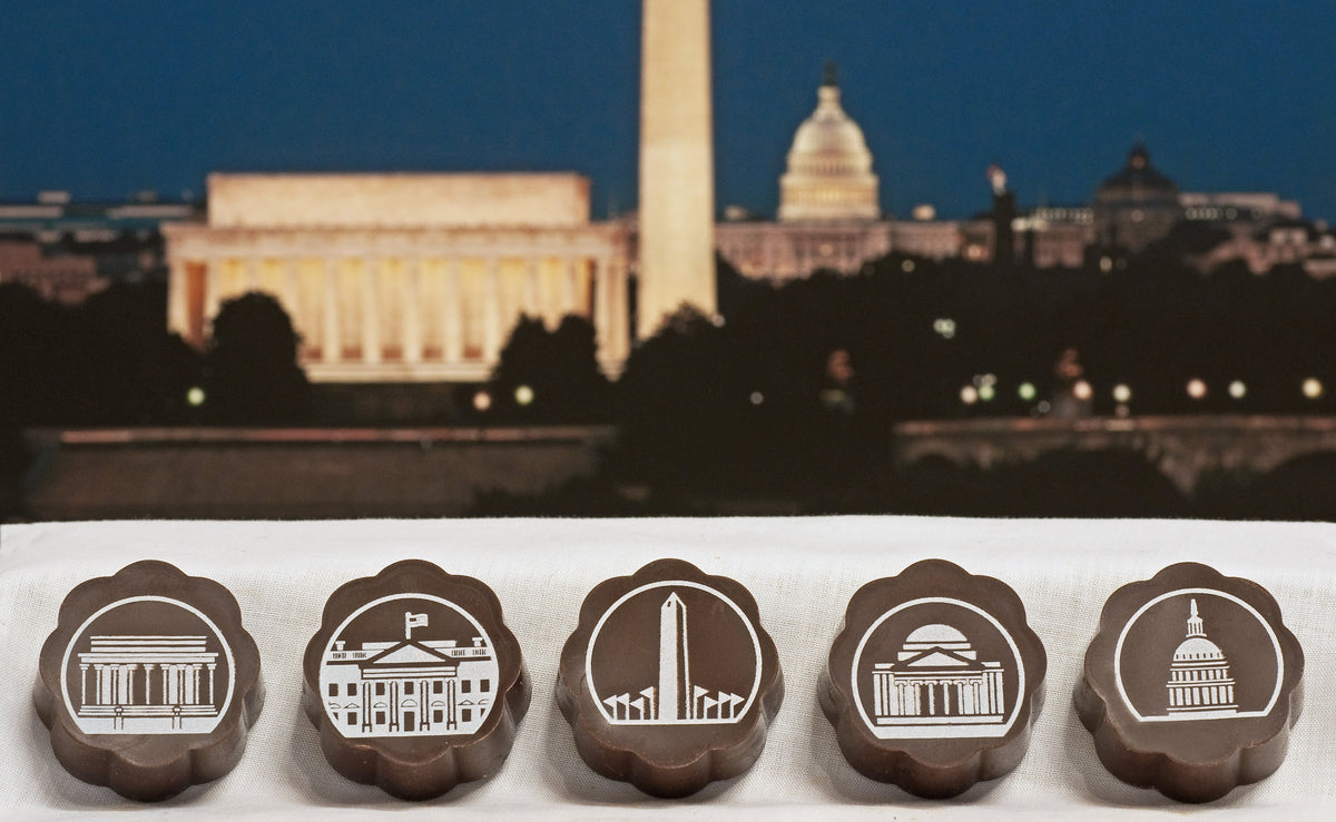 Five Chouquette Washington DC chocolates with white drawings of the Lincoln Memorial, White House, Washington Monument, Jefferson Memorial and Capitol, before a D.C. skyline