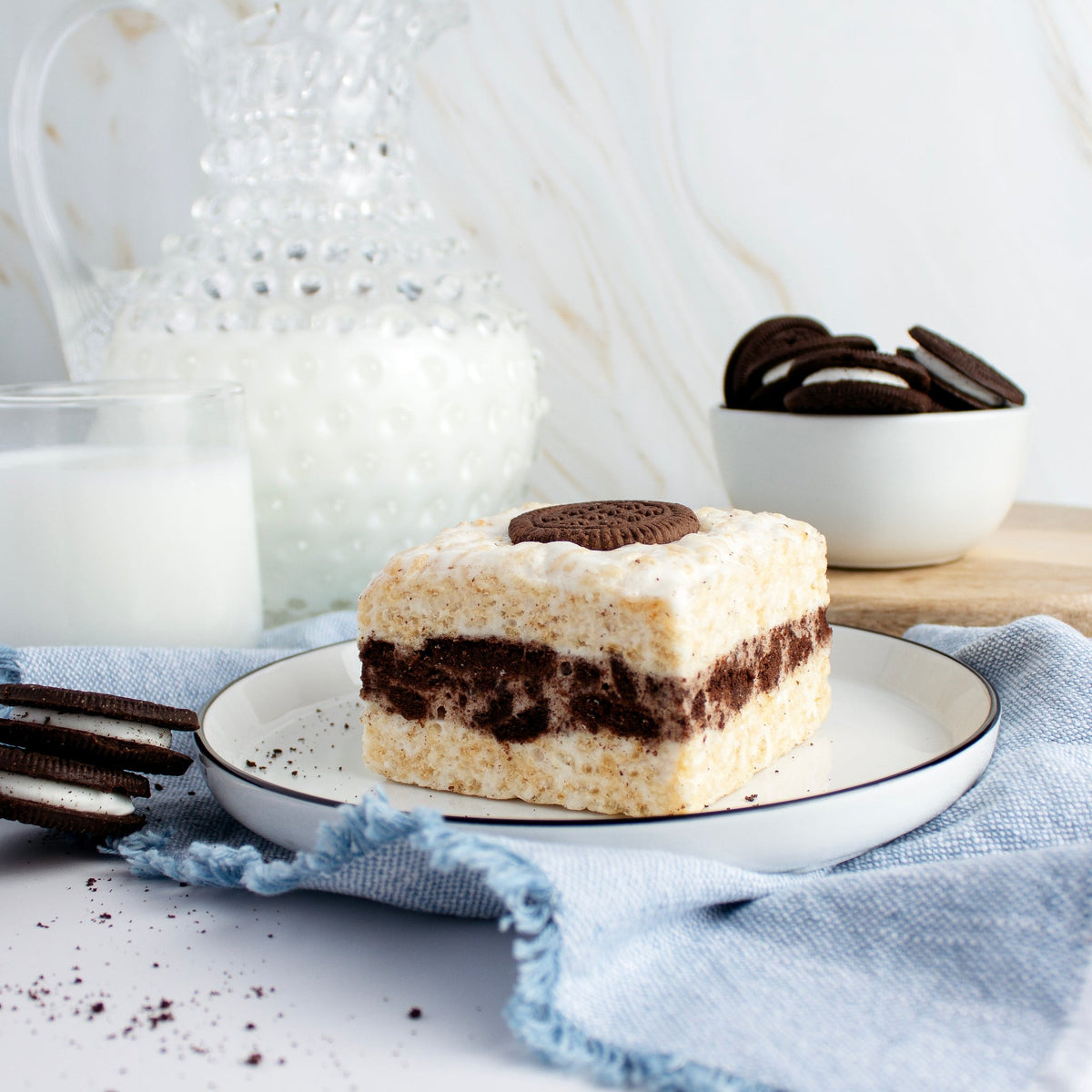 The Crispery Cookies & Cream Crispycake on a white plate beside a glass of milk and a bowl of chocolate sandwich cookies