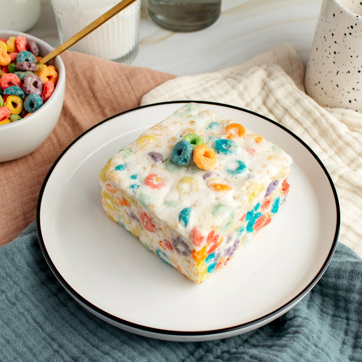 The Crispery Fruity Ringlets Crispycake on a white plate with a few cereal rings on top, beside a bowl of fruity ring cereal