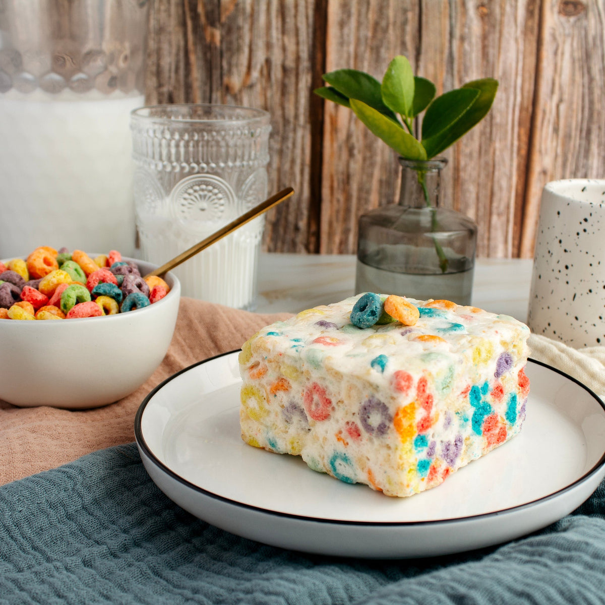 Side view of The Crispery Fruity Ringlets Crispycake on a plate, with a bowl of fruity cereal and a glass of milk behind it