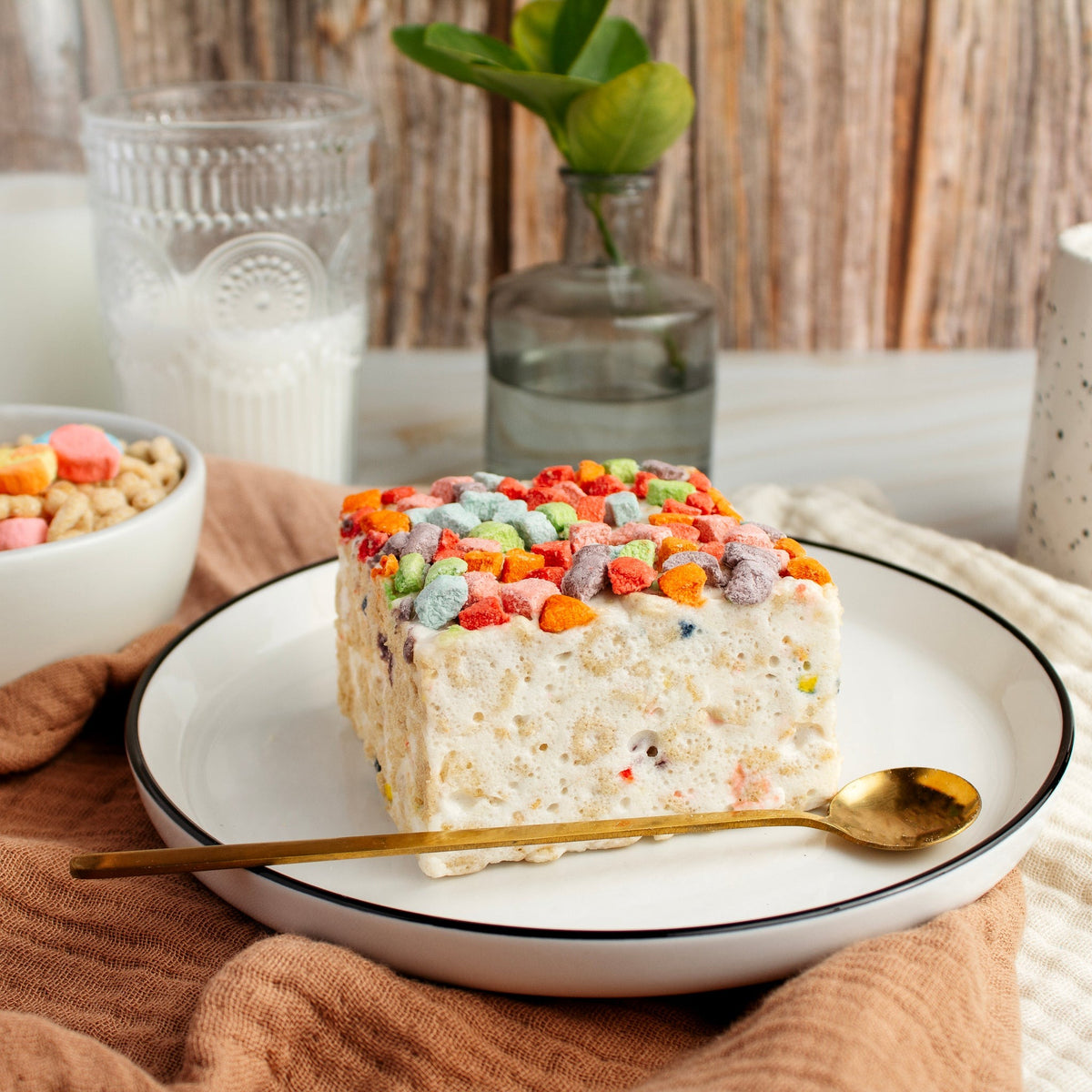 The Crispery Magical Charms Crispycake on a white plate with a gold spoon, beside a bowl of marshmallow cereal and a glass of milk