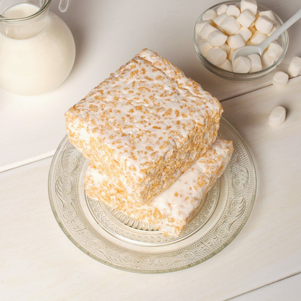 Two Plain & Sweet Crispycakes stacked on a glass plate beside a milk jug and a bowl of marshmallows