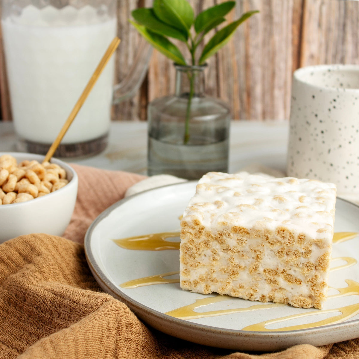 Honey Nut Crispy Crispycake on a plate with a honey drizzle, beside a bowl of oat O cereal and a glass of milk