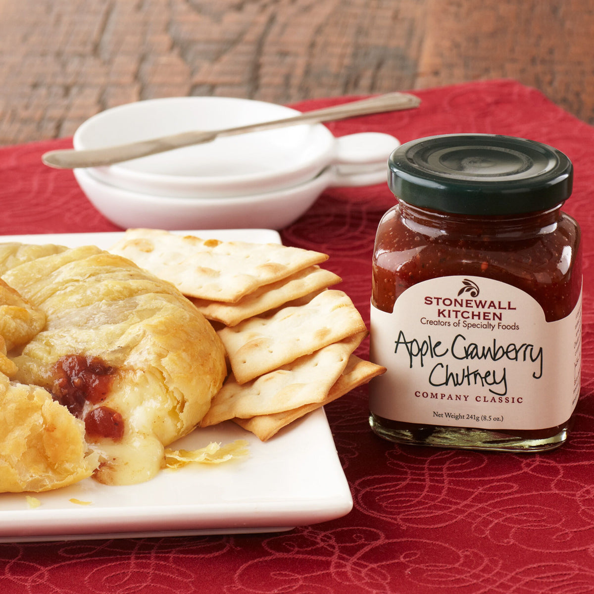 Stonewall Kitchen Apple Cranberry Chutney 8.5 oz jar beside baked brie in pastry and crackers on a red tablecloth