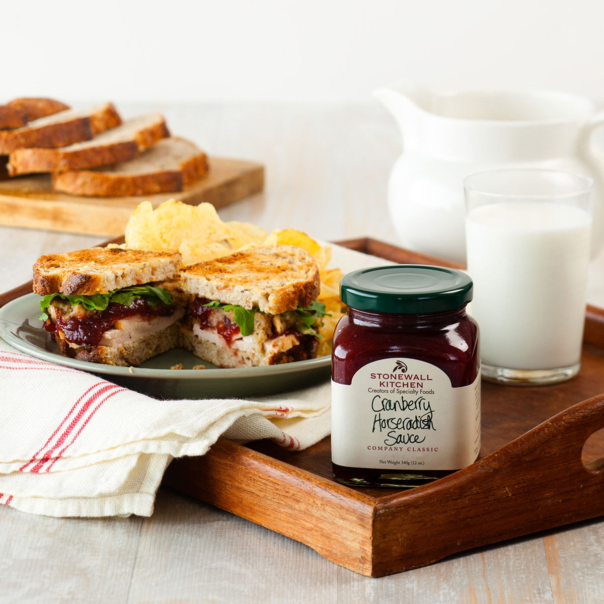 Jar of Stonewall Kitchen Cranberry Horseradish Sauce, 12 oz, on a wooden tray beside a grilled sandwich, chips and a glass of milk