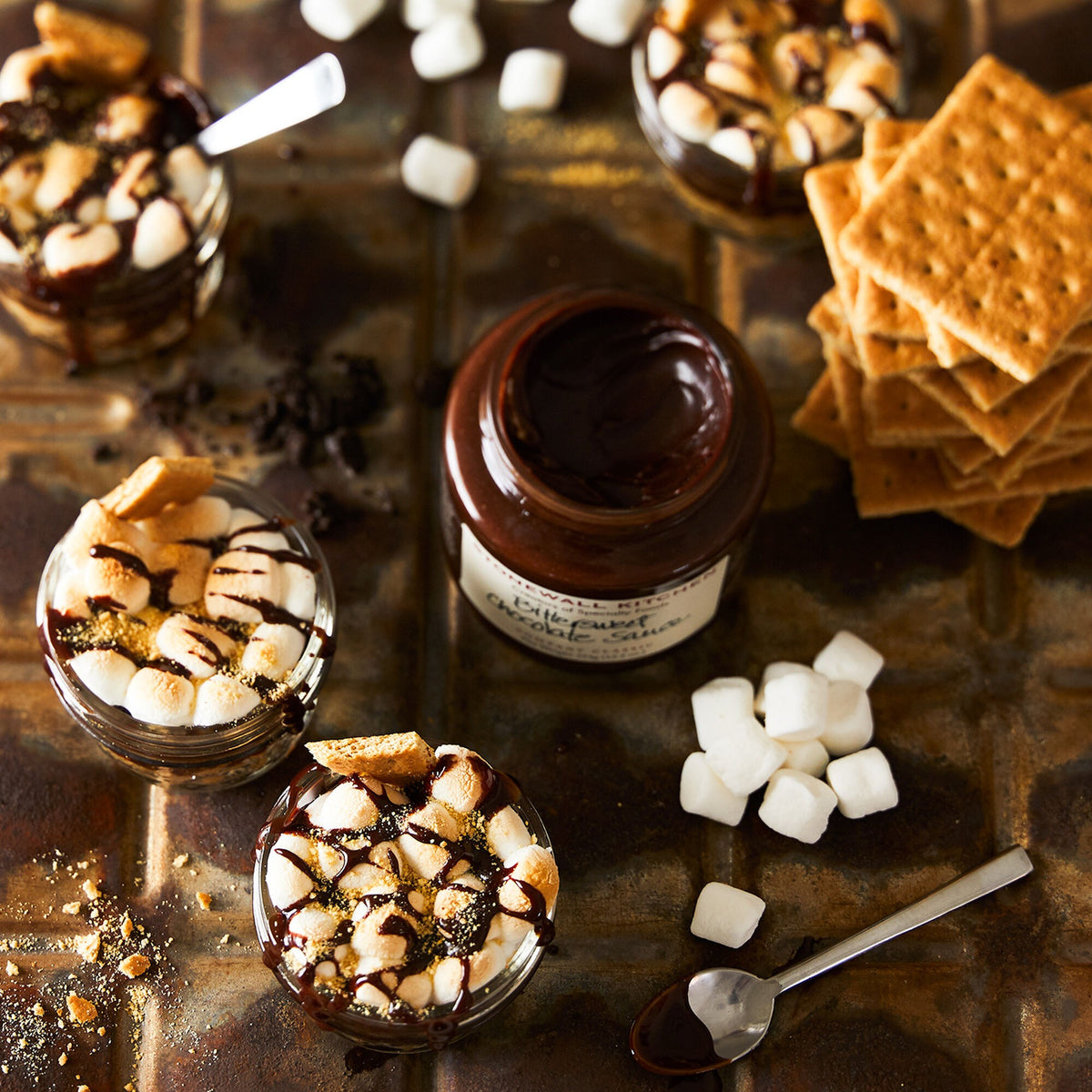 Open jar of Stonewall Kitchen Bittersweet Chocolate Sauce among s'mores dessert cups with toasted marshmallows and graham crackers