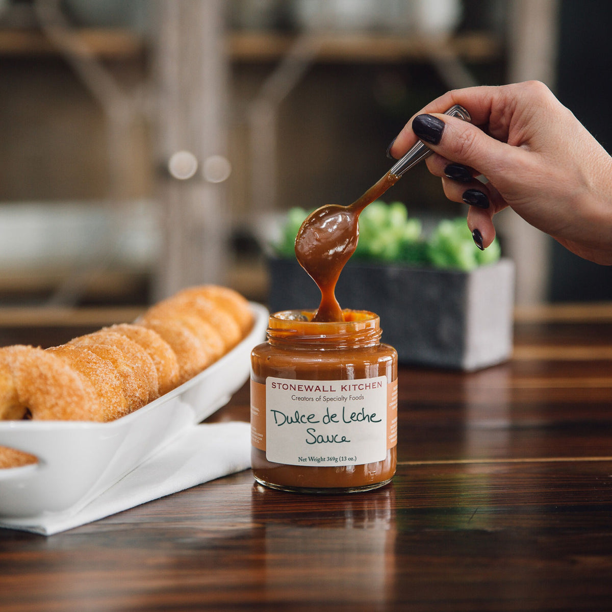 Spoon lifting Stonewall Kitchen Dulce De Leche Sauce from its jar on a wooden table next to a tray of donuts