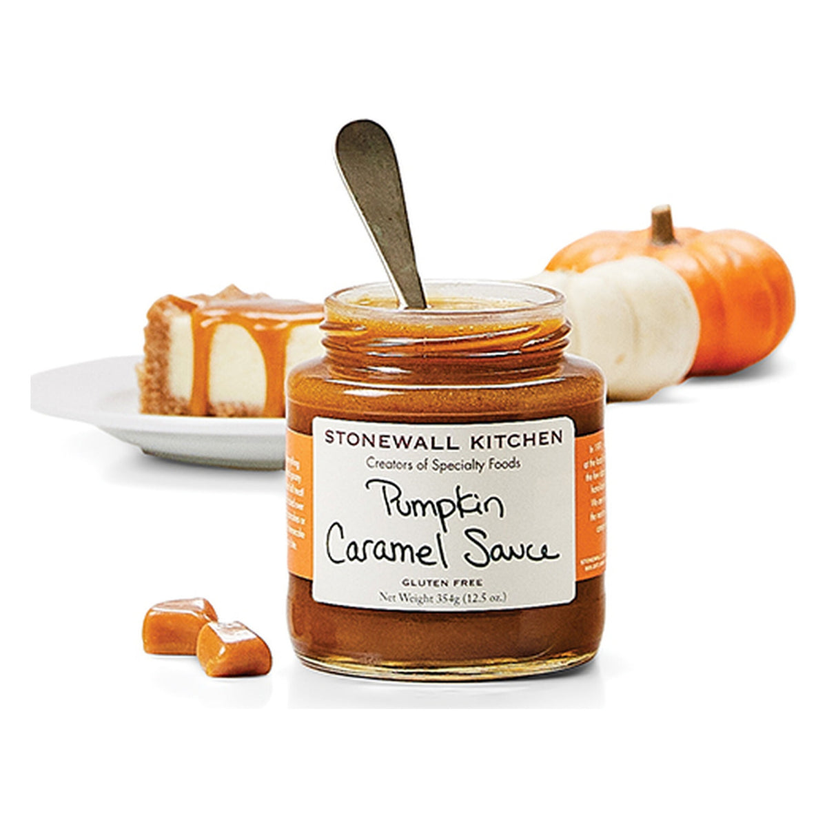 Open jar of Stonewall Kitchen Pumpkin Caramel Sauce with a spoon, beside a slice of caramel-topped cheesecake, caramels and small pumpkins