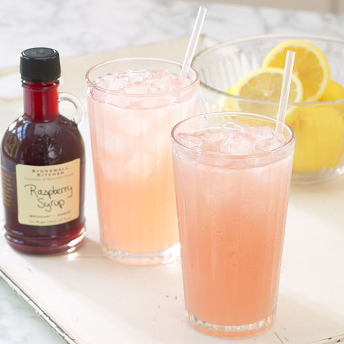 Bottle of Stonewall Kitchen Raspberry Syrup in an earlier label design beside two glasses of raspberry lemonade and a bowl of lemons